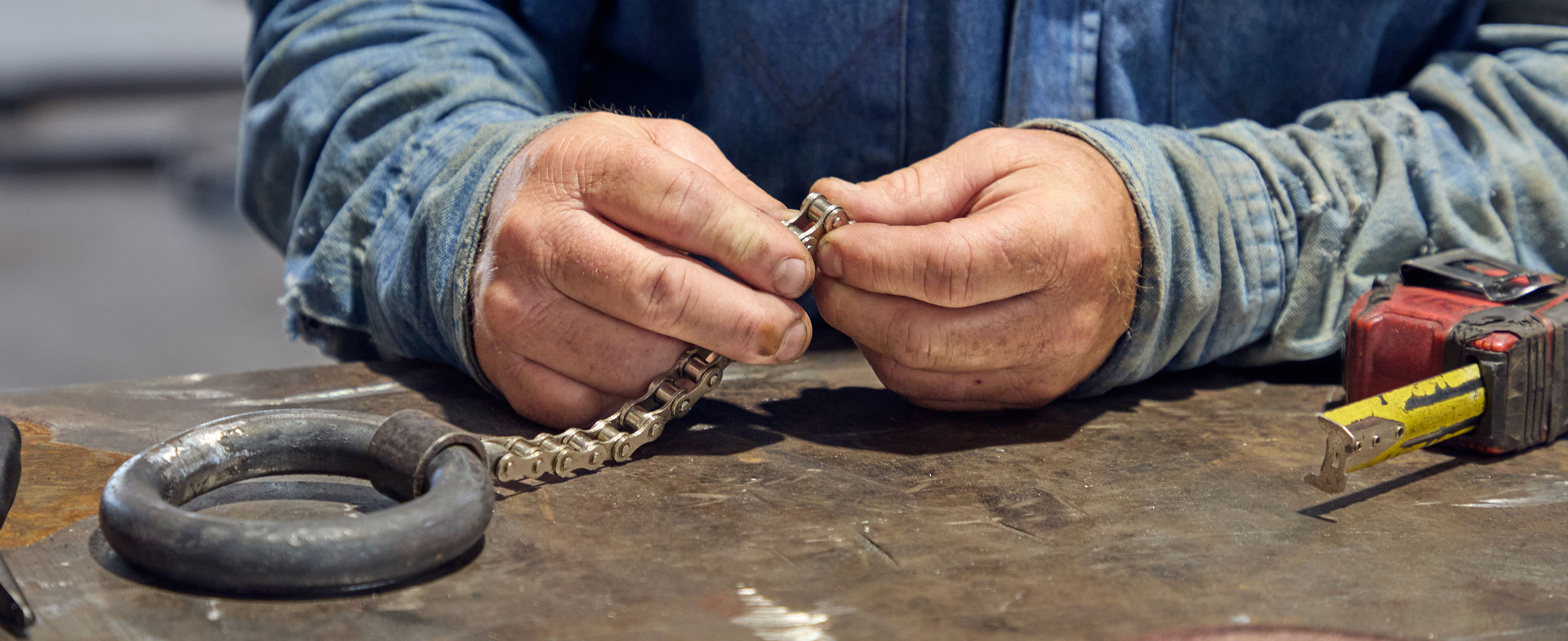 Primer plano de un hombre con camisa vaquera trabajando con una cadena metálica en una mesa de taller, rodeado de herramientas como una cinta métrica y un grillete.